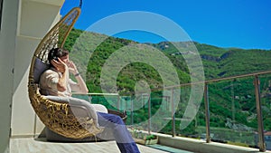 A young man on a balcony is enjoying music in a swinging chair. Mountain scenery in the background. His headphones are