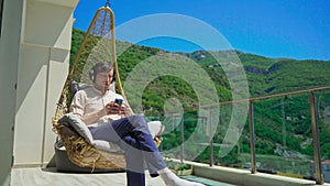 A young man on a balcony is enjoying music in a swinging chair. Mountain scenery in the background. His headphones are