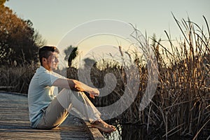 Young man on bare feet sitting on wooden walkway on lake shore during sunset. Inspirational concept