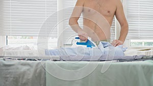 Young man with bare torso standing behind board using steaming iron, ironing shirt during housework. 4k, slow-motion