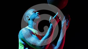 Young man, basketball player spinning basketball ball on his finger at studio over dark background in neon. Sport