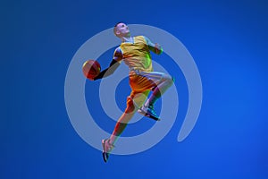 Young man, basketball player in yellow uniform training, throwing ball in a jump against blue studio background in neon