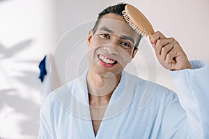 Young man in bathrobe combing hair with hairbrush while amiling and looking at camera