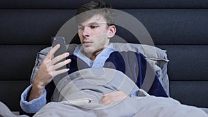 Young man in bathrobe lying in bed and reading news on smartphone before sleep. Male lies in bed and looking at phone screen in th