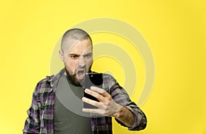 A young man with a beard and a bald head holding a phone in his hands on a yellow background. A man beside himself with anger