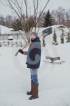 A young man with a beard in a blue jacket cleans snow with a shovel in his area 1