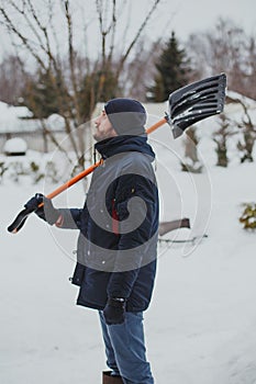 A young man with a beard in a blue jacket cleans snow with a shovel in his area 1