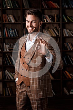 young man with a beard in a brown suit shows ok gesture standing in the library. The businessman is smiling and pleased
