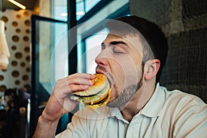 A young man with a beard eating a burger at the restaurant and enjoying the taste