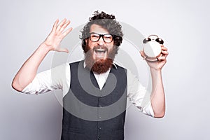 Young man with beard and glasses is holding a clock and screaming is looking at the camera