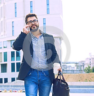 Young man with beard in glasses holding coffee cup and talking on mobile phone while walking outdoors.