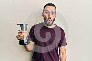 Young man with beard holding coffee maker with filter scared and amazed with open mouth for surprise, disbelief face