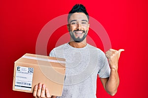 Young man with beard holding delivery package pointing thumb up to the side smiling happy with open mouth