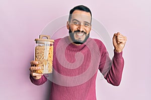 Young man with beard holding jar with macaroni pasta screaming proud, celebrating victory and success very excited with raised arm