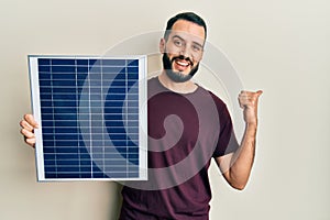 Young man with beard holding photovoltaic solar panel pointing thumb up to the side smiling happy with open mouth