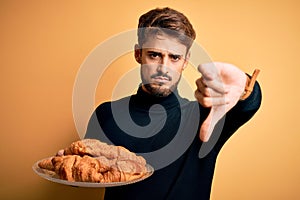 Young man with beard holding plate with croissants standing over isolated yellow background with angry face, negative sign showing