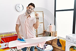 Young man with beard ironing clothes at home pointing to the back behind with hand and thumbs up, smiling confident