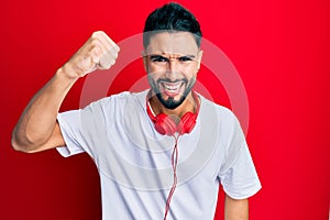 Young man with beard listening to music using headphones angry and mad raising fist frustrated and furious while shouting with