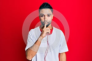 Young man with beard listening to music using headphones asking to be quiet with finger on lips