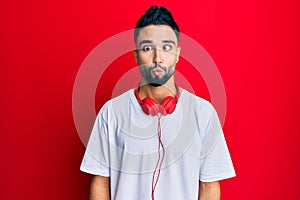 Young man with beard listening to music using headphones making fish face with lips, crazy and comical gesture