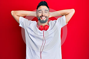 Young man with beard listening to music using headphones relaxing and stretching, arms and hands behind head and neck smiling