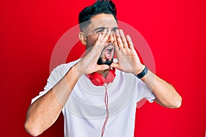 Young man with beard listening to music using headphones shouting angry out loud with hands over mouth