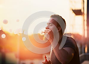 Young man with a beard makes the bubble with the steam inside, creating soft background of the urban landscape