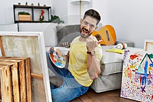 Young man with beard painting canvas at home pointing to the back behind with hand and thumbs up, smiling confident