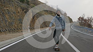 Young man with a beard riding skateboard cruising downhill on countryside road