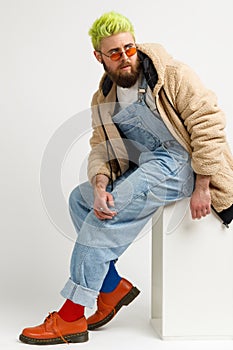 Young man with beard and trendy green hair wearing stylish clothing and shoes, sitting on white cube and looking away with pensive