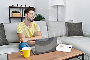 Young man with beard using laptop at home looking away to side with smile on face, natural expression