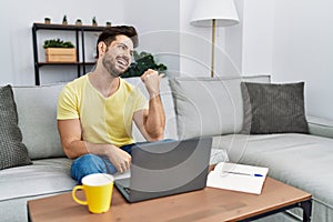 Young man with beard using laptop at home smiling with happy face looking and pointing to the side with thumb up