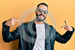 Young man with beard wearing black leather jacket and sunglasses looking confident with smile on face, pointing oneself with