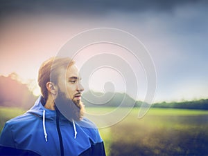 Young man with a beard wearing a blue jacket and in profile against green field and sky