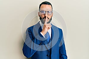 Young man with beard wearing business suit and tie asking to be quiet with finger on lips