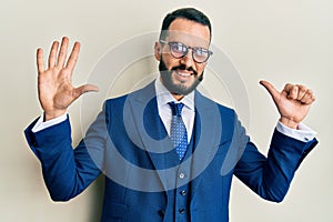 Young man with beard wearing business suit and tie showing and pointing up with fingers number six while smiling confident and