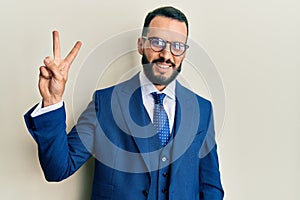 Young man with beard wearing business suit and tie showing and pointing up with fingers number two while smiling confident and