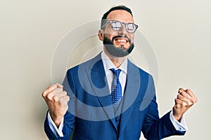 Young man with beard wearing business suit and tie very happy and excited doing winner gesture with arms raised, smiling and