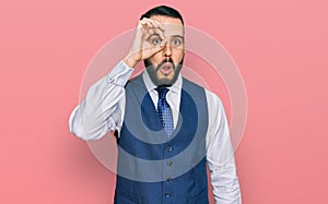 Young man with beard wearing business vest doing ok gesture shocked with surprised face, eye looking through fingers