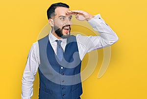 Young man with beard wearing business vest very happy and smiling looking far away with hand over head
