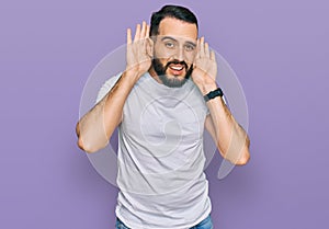 Young man with beard wearing casual white t shirt trying to hear both hands on ear gesture, curious for gossip