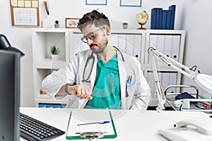 Young man with beard wearing doctor uniform and stethoscope at the clinic checking the time on wrist watch, relaxed and confident