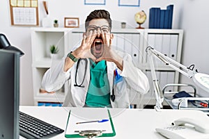 Young man with beard wearing doctor uniform and stethoscope at the clinic shouting angry out loud with hands over mouth