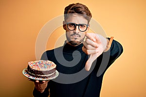 Young man with beard wearing glasses holding birthday cake over isolated yellow background with angry face, negative sign showing