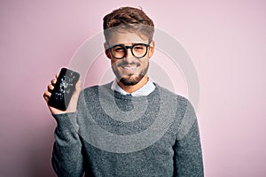 Young man with beard wearing glasses holding broken and craked smartphone with a happy face standing and smiling with a confident