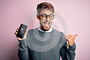 Young man with beard wearing glasses holding broken and craked smartphone pointing and showing with thumb up to the side with