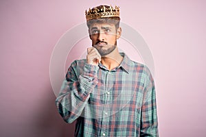 Young man with beard wearing golden crown of king standing over isolated pink background with hand on chin thinking about