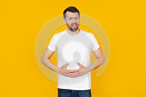 A young man with a beard in a white T-shirt with a hand on his stomach because of indigestion, feeling sick. Pain concept. Stands