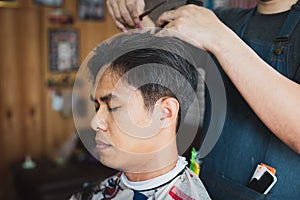 Young man being haircut with scissor by professional barber in barbershop. Hairdresser using comb and scissors cut hair