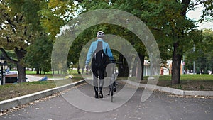 A young man in a bicycle walks through the park. Goes down the descent
